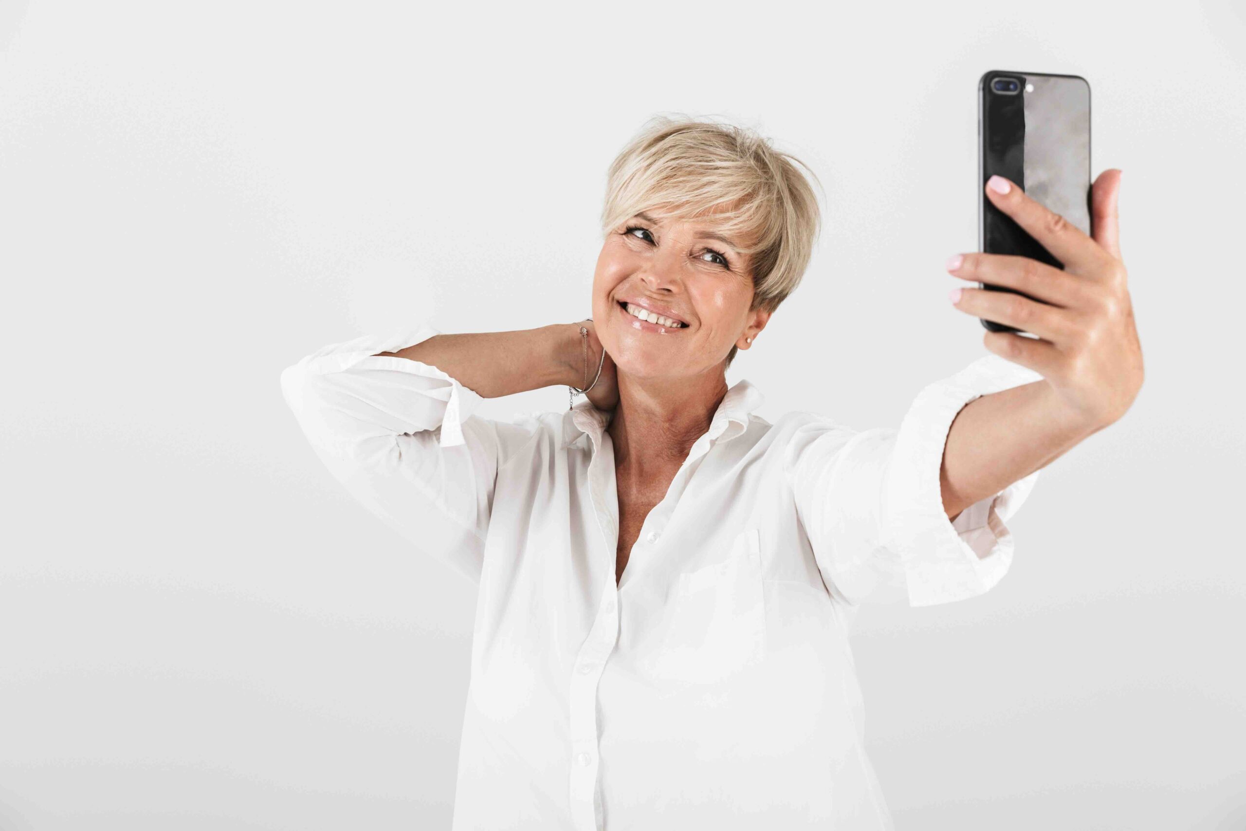 Image of joyous adult woman with short blond hair smiling and taking selfie photo on cellphone isolated over white background in studio